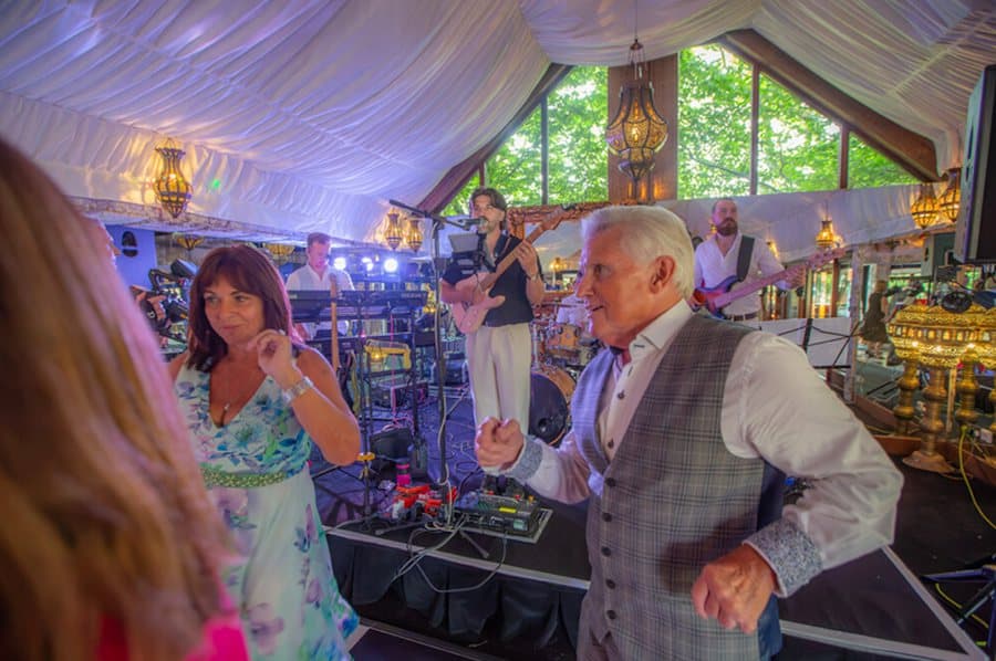 Guests dancing in front of the band under a marquee, lanterns overhead and daylight through the windows